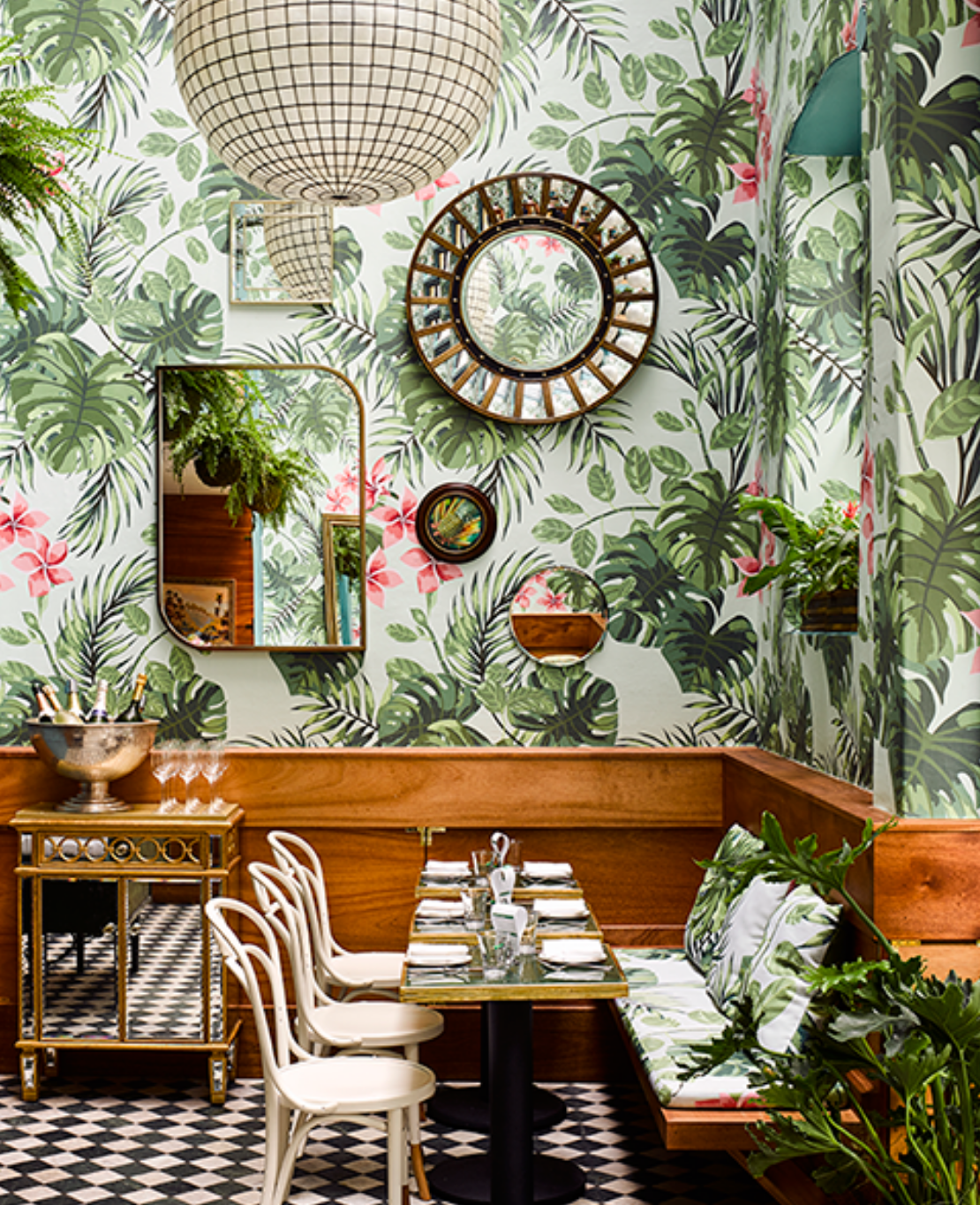 A vibrant restaurant corner with tropical wallpaper, wood paneling, a checkered floor, and stylish mirrors, featuring a set dining table and hanging plants.